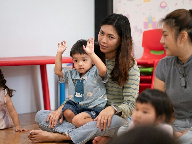 Guru memangku balita yang mengangkat kedua tangannya di kelas