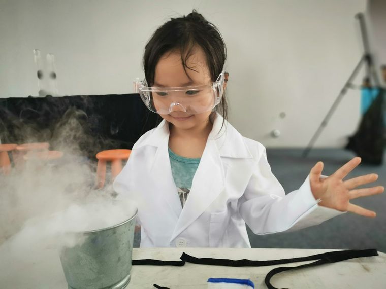 Anak perempuan berjas lab dan kacamata pelindung melihat asap dari es kering